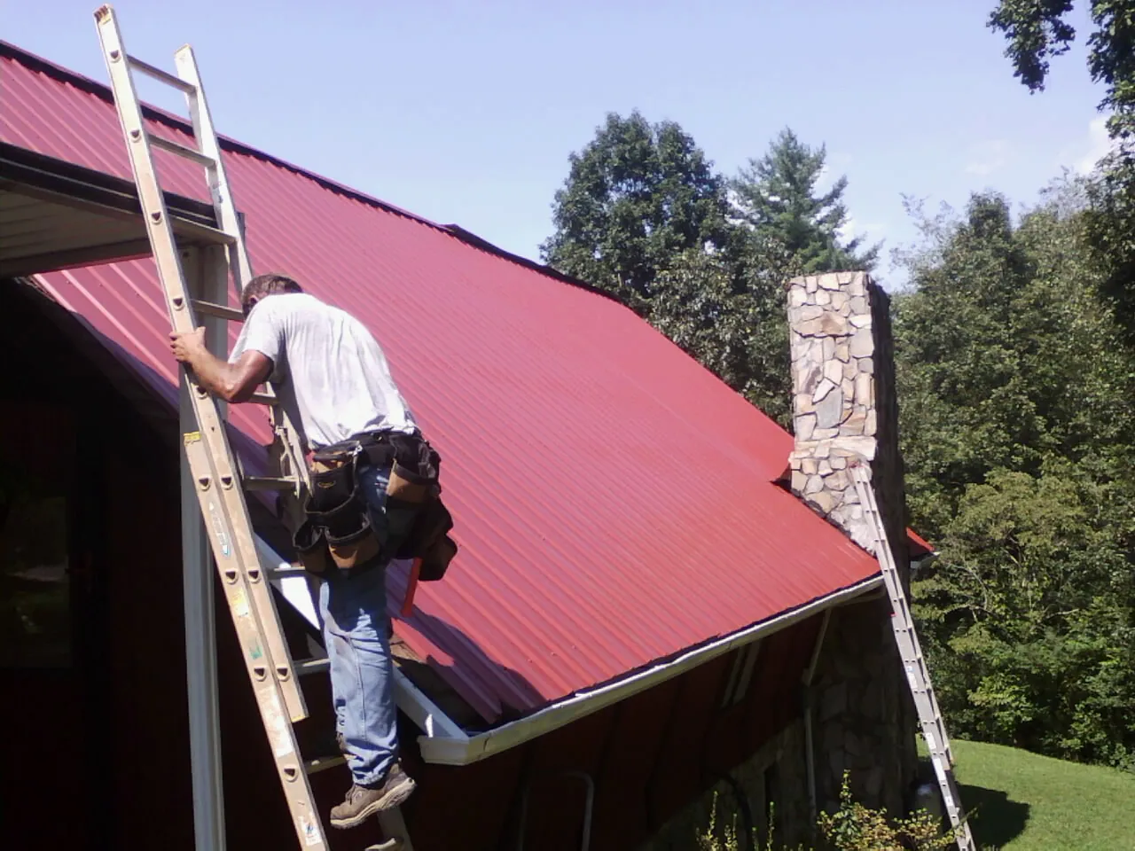 Roofer on a ladder beside a red metal roof and stone chimney — Soffit & Fascia Repair by Roofing Brothers in Edgar